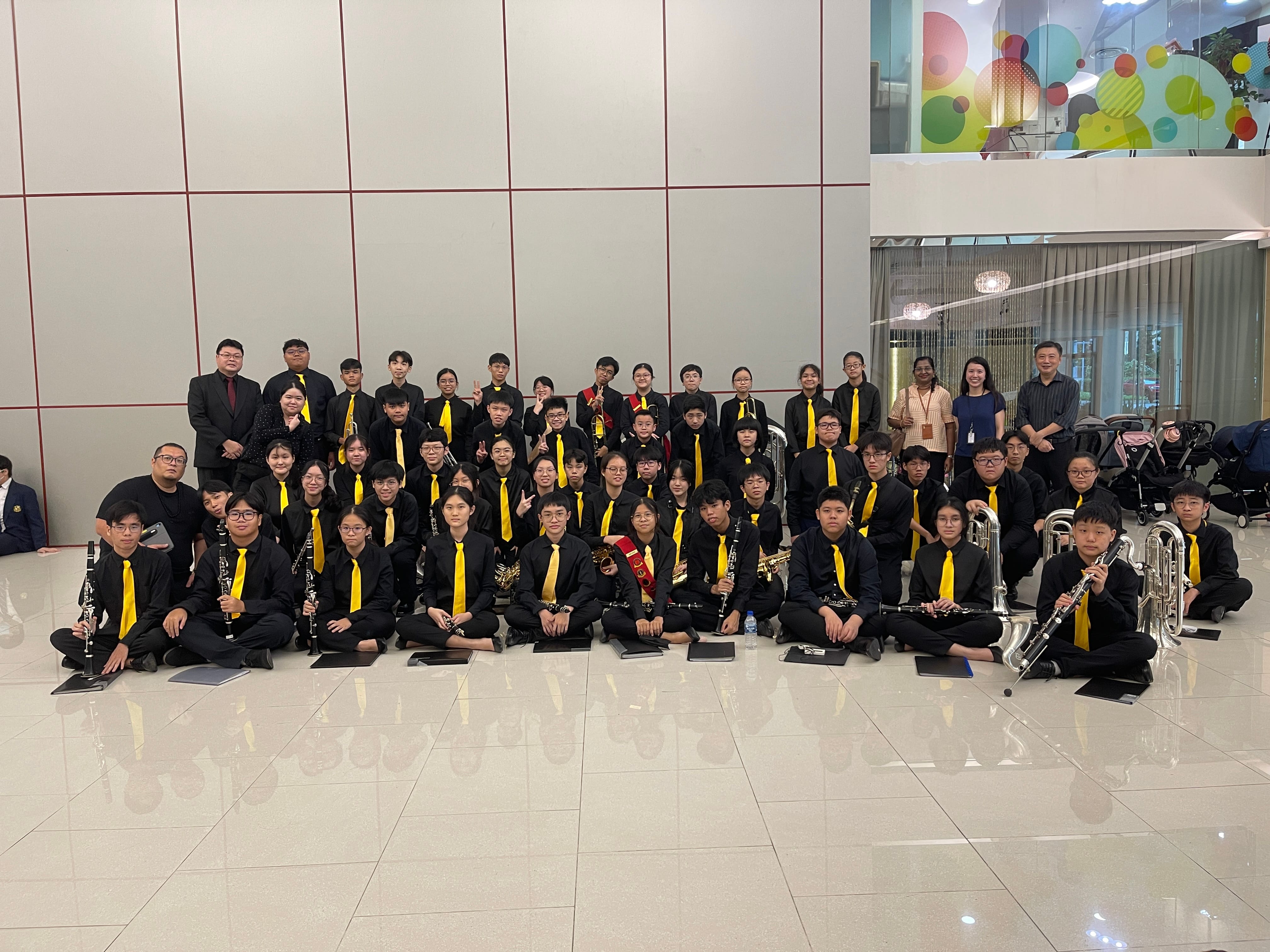 NV Symphonic Band members in black uniforms with yellow ties posing indoors, holding clarinets and brass instruments.